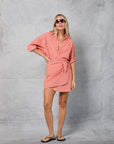 A woman in sunglasses, a coral and white striped shirt dress tied at the waist, black flip-flops, and the Mamene Repeat Wrap Tennis Skirt stands before a gray textured backdrop, creating a playful warm-weather vibe.