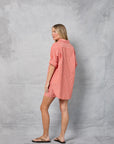 A woman stands sideways, looking back, wearing Mamene's Repeat Shirt— a loose-fitting coral and white Japanese yarn-dye cotton poplin top—paired with matching shorts and black flip-flops against a light gray textured backdrop.