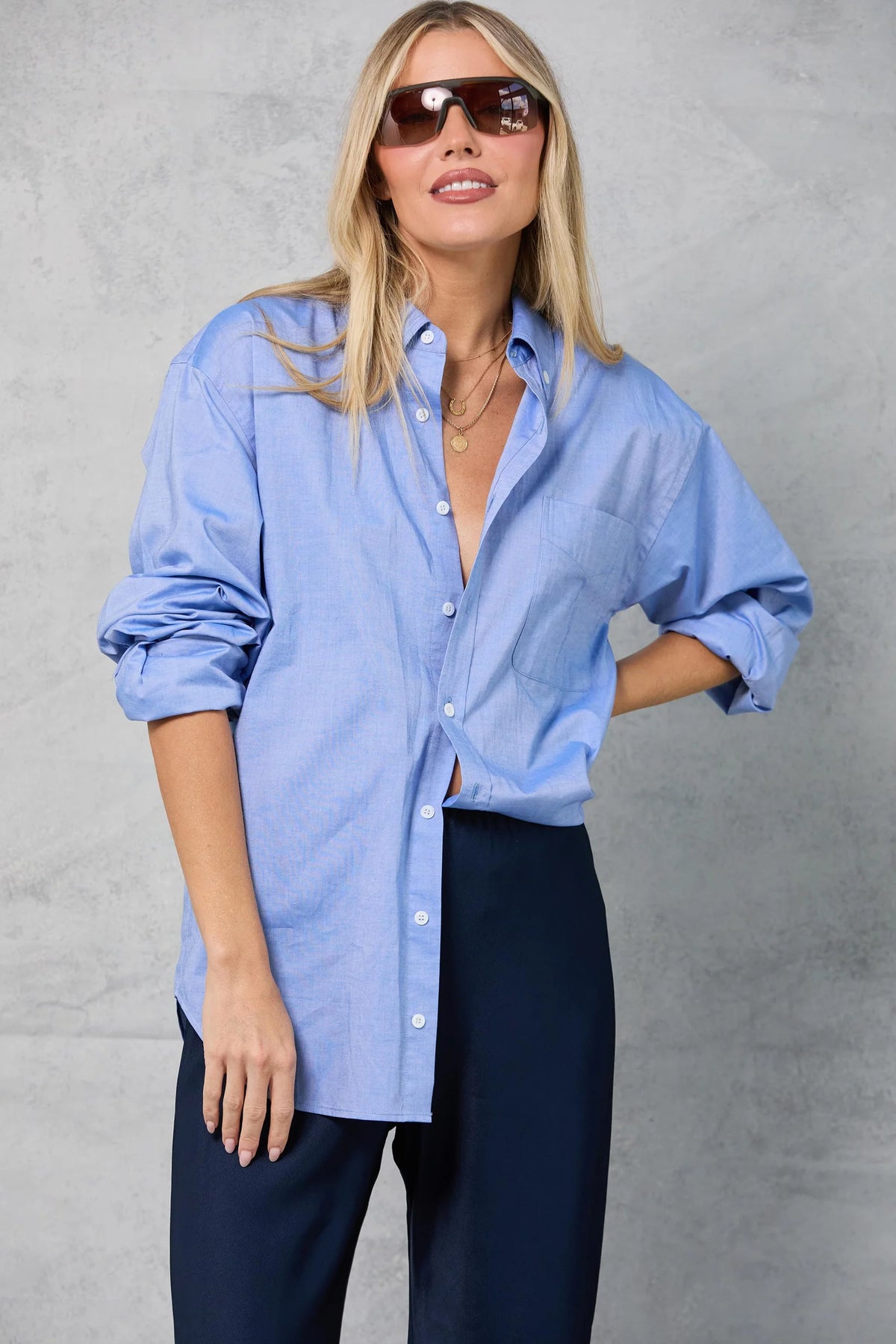 A woman wearing sunglasses and Mamene’s Slim Repeat Shirt in light blue cotton poplin with rolled sleeves, paired with dark trousers, stands against a light gray textured background, smiling slightly with one hand in her pocket.