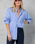 A woman wearing sunglasses and Mamene’s Slim Repeat Shirt in light blue cotton poplin with rolled sleeves, paired with dark trousers, stands against a light gray textured background, smiling slightly with one hand in her pocket.