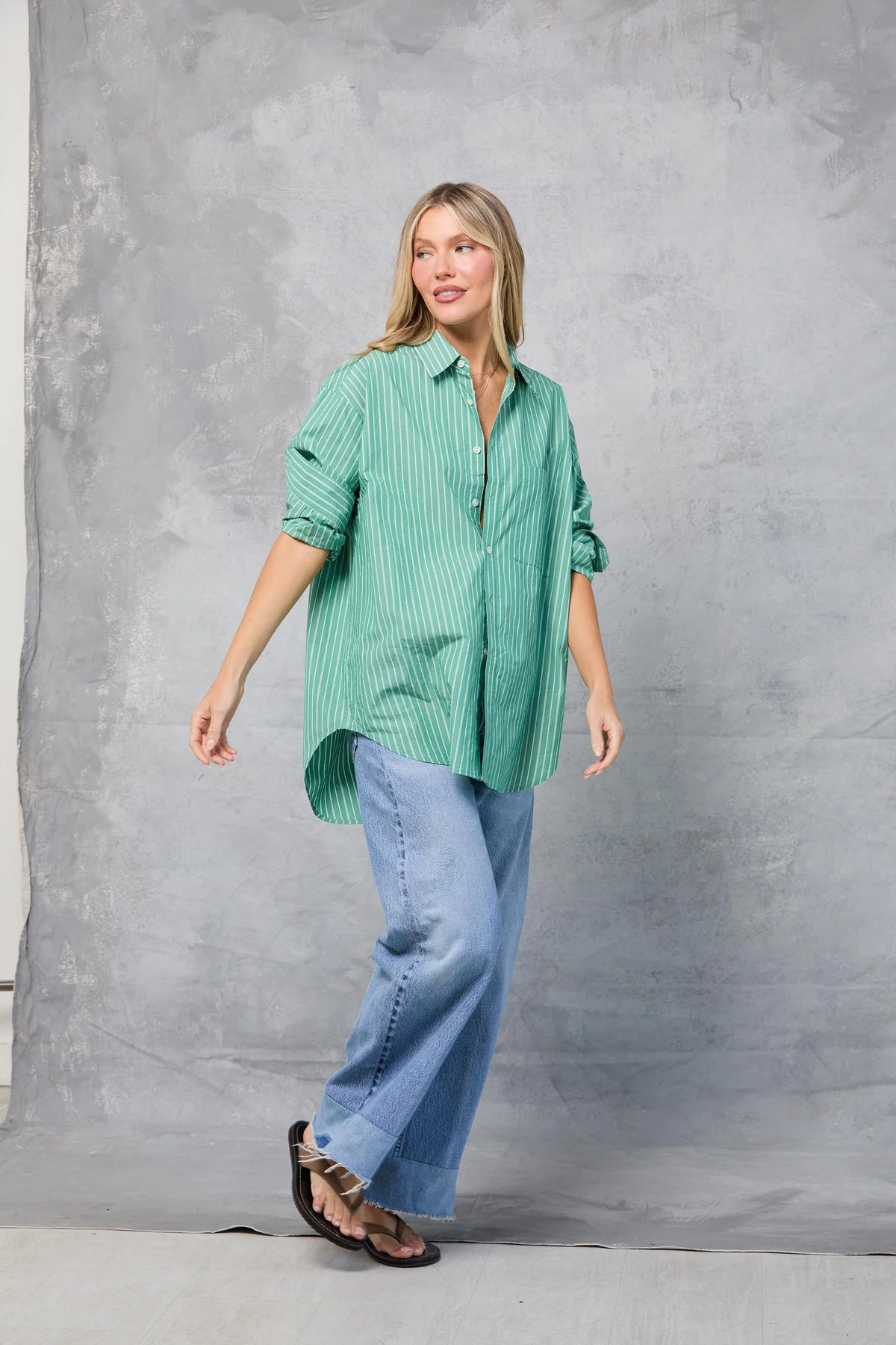 A woman stands before a gray textured backdrop, smiling and looking to the side. She wears Mamene's Repeat Shirt in green and white with light blue wide-leg jeans and black sandals.