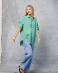 A woman stands before a gray textured backdrop, smiling and looking to the side. She wears Mamene's Repeat Shirt in green and white with light blue wide-leg jeans and black sandals.