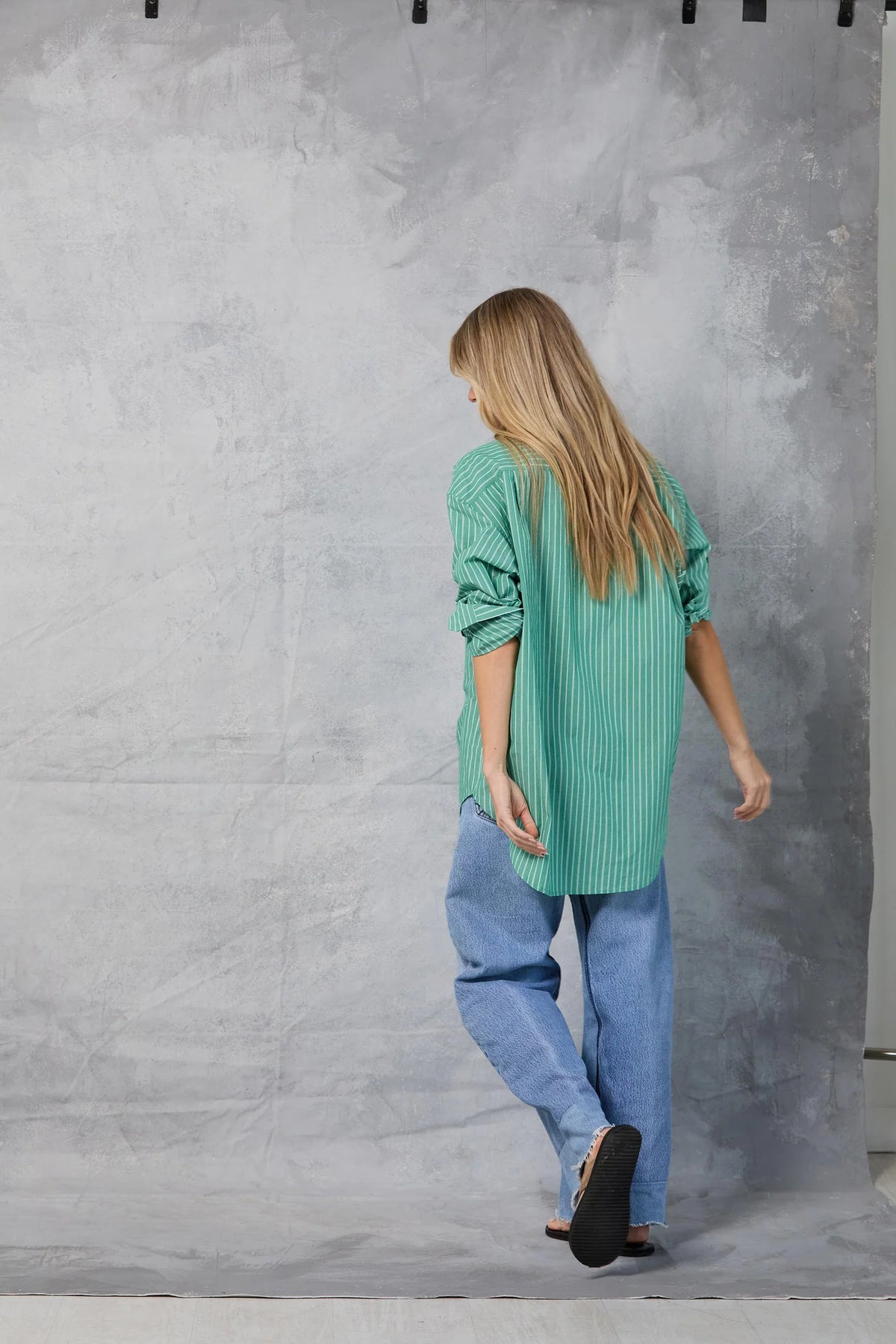 A person with long blonde hair, barefoot and facing away from the camera, models Mamene’s Repeat Shirt—a loose green striped cotton poplin top—paired with blue jeans against a gray textured backdrop.
