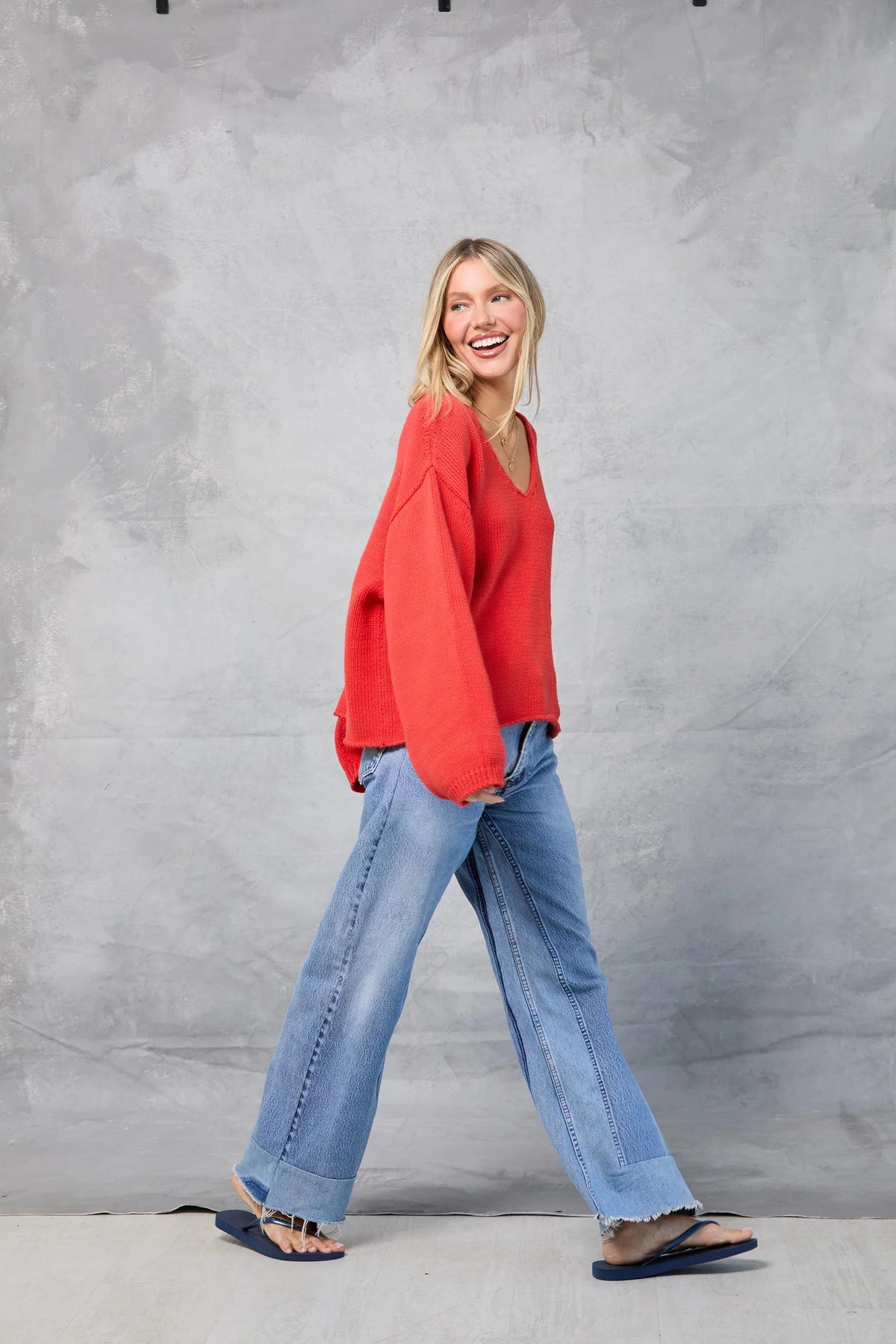 A smiling woman with blonde hair glances left as she walks in front of a gray textured backdrop, wearing the Mamene Winona Sweater in red, paired with blue jeans and flip-flops.