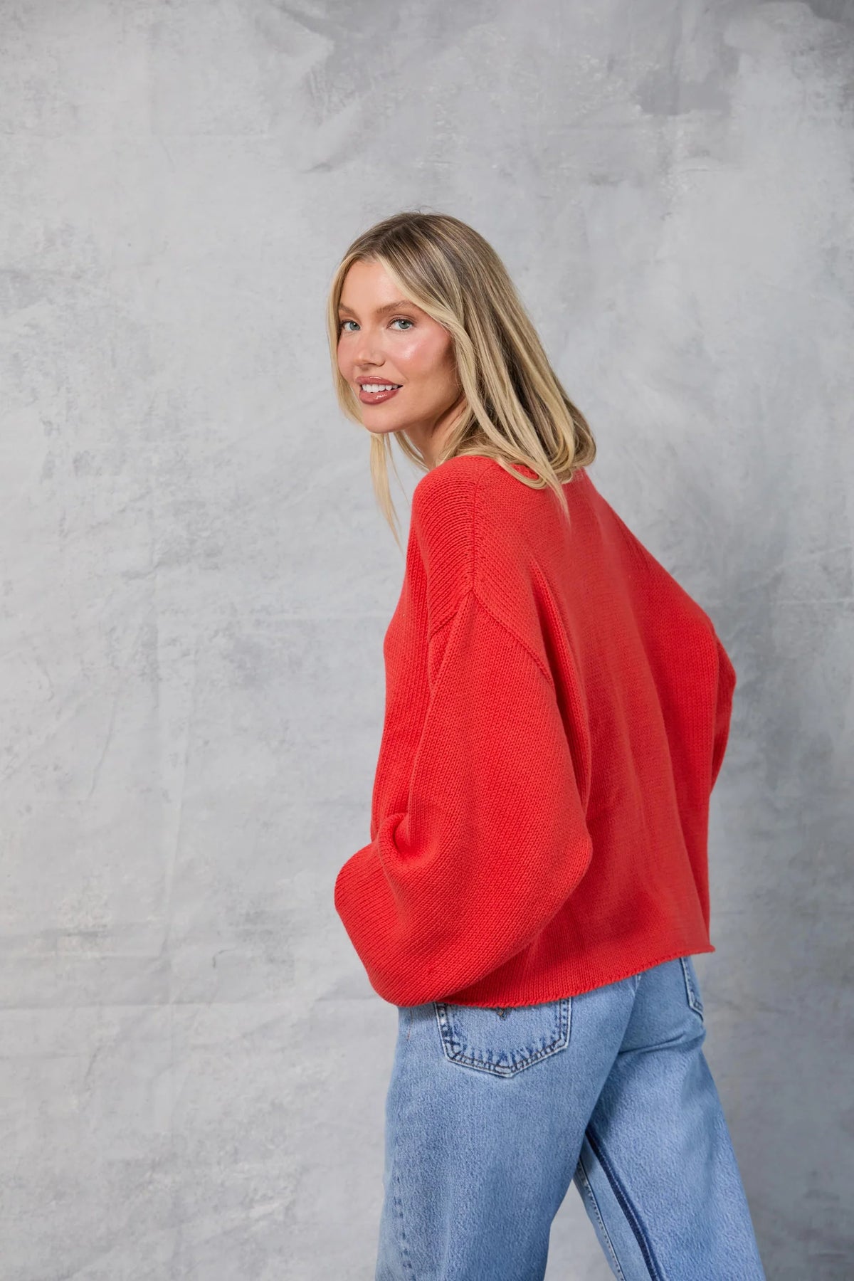 A woman with straight blonde hair stands sideways against a light gray textured backdrop, smiling over her shoulder while wearing the loose, oversized red Mamene Winona Sweater and blue jeans with her hands in her pockets.
