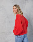 A woman with straight blonde hair stands sideways against a light gray textured backdrop, smiling over her shoulder while wearing the loose, oversized red Mamene Winona Sweater and blue jeans with her hands in her pockets.