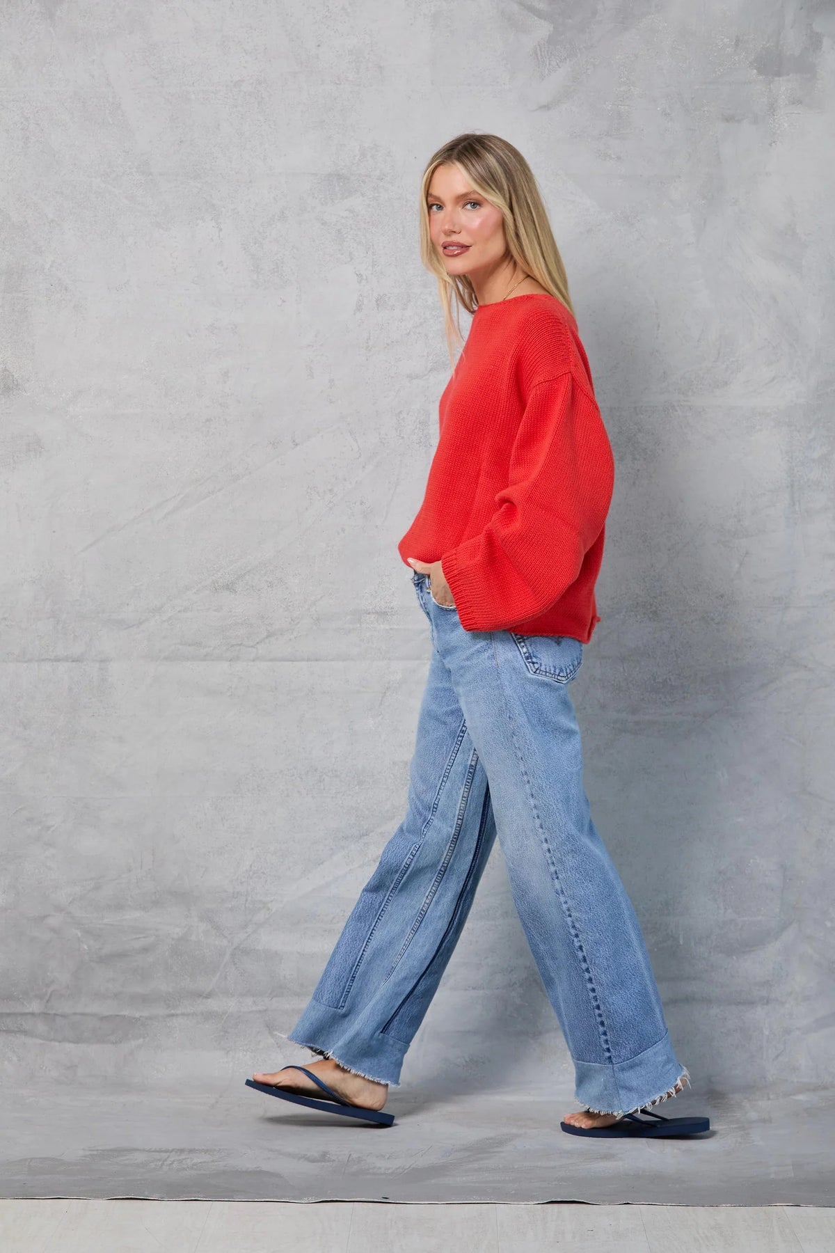 A woman with long blonde hair wears the Mamene Winona Sweater, an oversized bright red knit, paired with wide-leg blue jeans and flip-flops as she walks in front of a gray textured background.