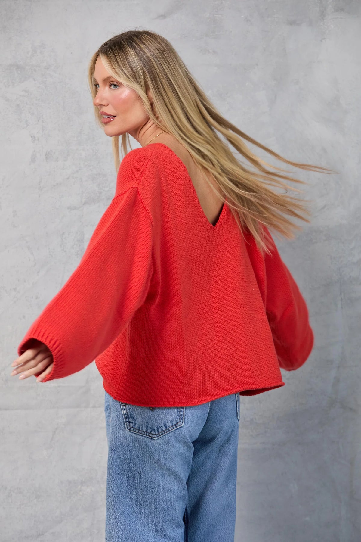 A woman with long blonde hair models the Mamene Winona Sweater, an oversized red cotton knit with a V-shaped back, paired with blue jeans as she poses sideways in front of a light gray textured wall.
