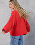 A woman with long blonde hair models the Mamene Winona Sweater, an oversized red cotton knit with a V-shaped back, paired with blue jeans as she poses sideways in front of a light gray textured wall.