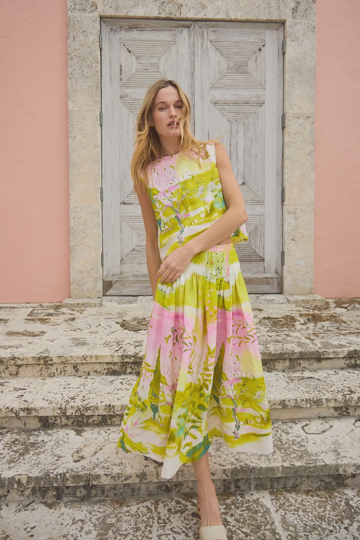 A person stands on stone steps in front of a textured wooden door and pink wall, wearing the vibrant Fallon Skirt by Hunter Bell NYC with custom print patterns in green, pink, and white, creating a summery and stylish atmosphere.