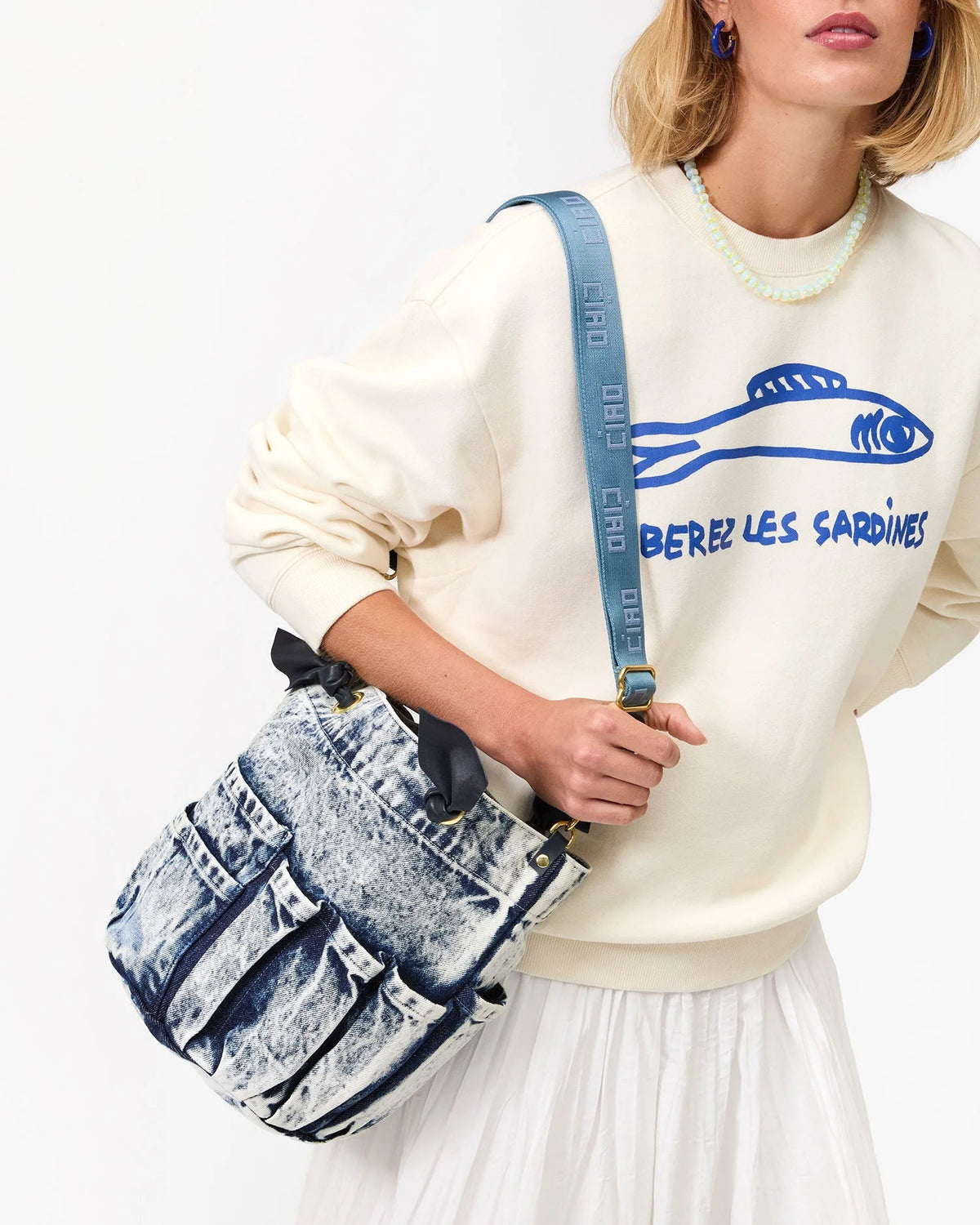 A woman wears a cream sweatshirt with a blue fish graphic and "LIBEREZ LES SARDINES" text, a white pleated skirt, and carries a blue denim handbag featuring the Clare Vivier Adjustable Crossbody Strap over her shoulder.