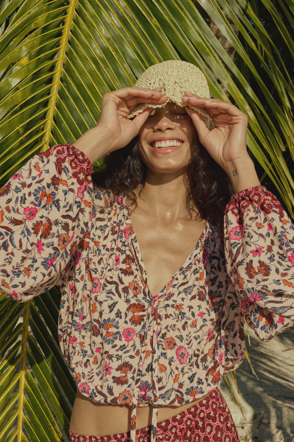 A smiling person stands in sunlight before large palm leaves, wearing the colorful Amelia Top by natalie martin and lifting a woven hat above their eyes.