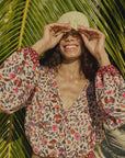 A smiling person stands in sunlight before large palm leaves, wearing the colorful Amelia Top by natalie martin and lifting a woven hat above their eyes.
