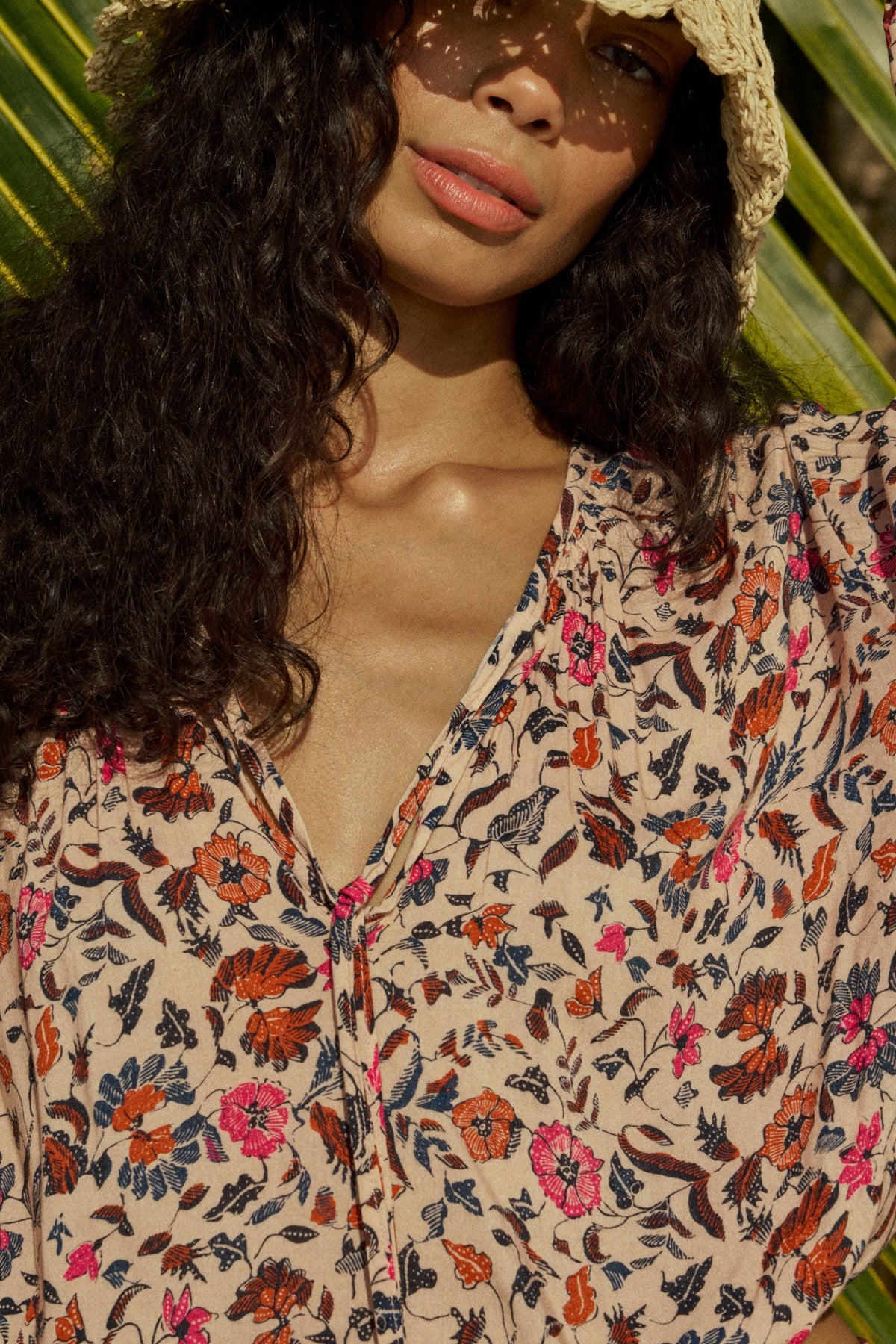A woman with long curly hair wears the natalie martin Amelia Top, a floral v-neck blouse in red, pink, and blue, paired with a wide-brim straw hat, standing in sunlight among palm leaves.