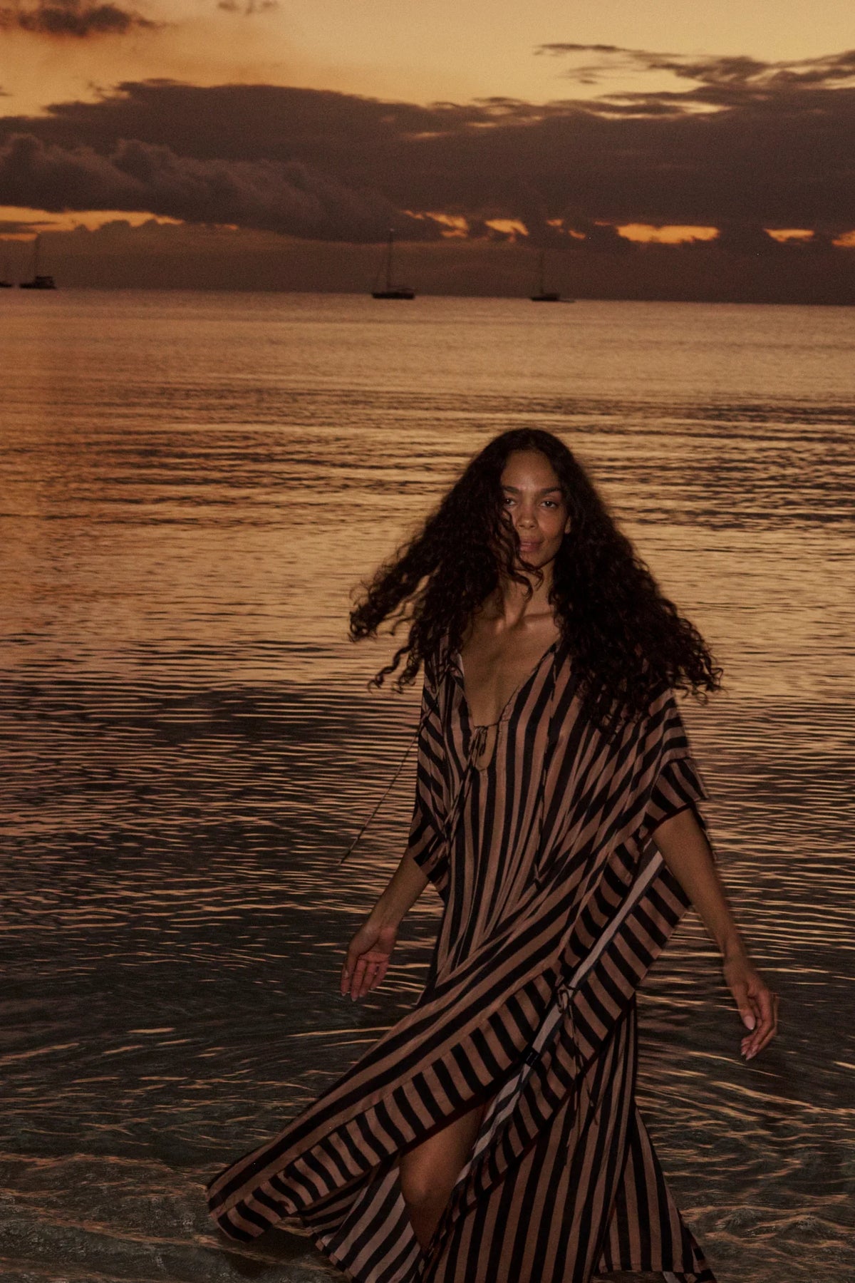 Paulina Caftan by Natalie Martin in a sunset coastal setting, featuring soft clouds and anchored sailboats.