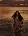 Paulina Caftan by Natalie Martin in a sunset coastal setting, featuring soft clouds and anchored sailboats.