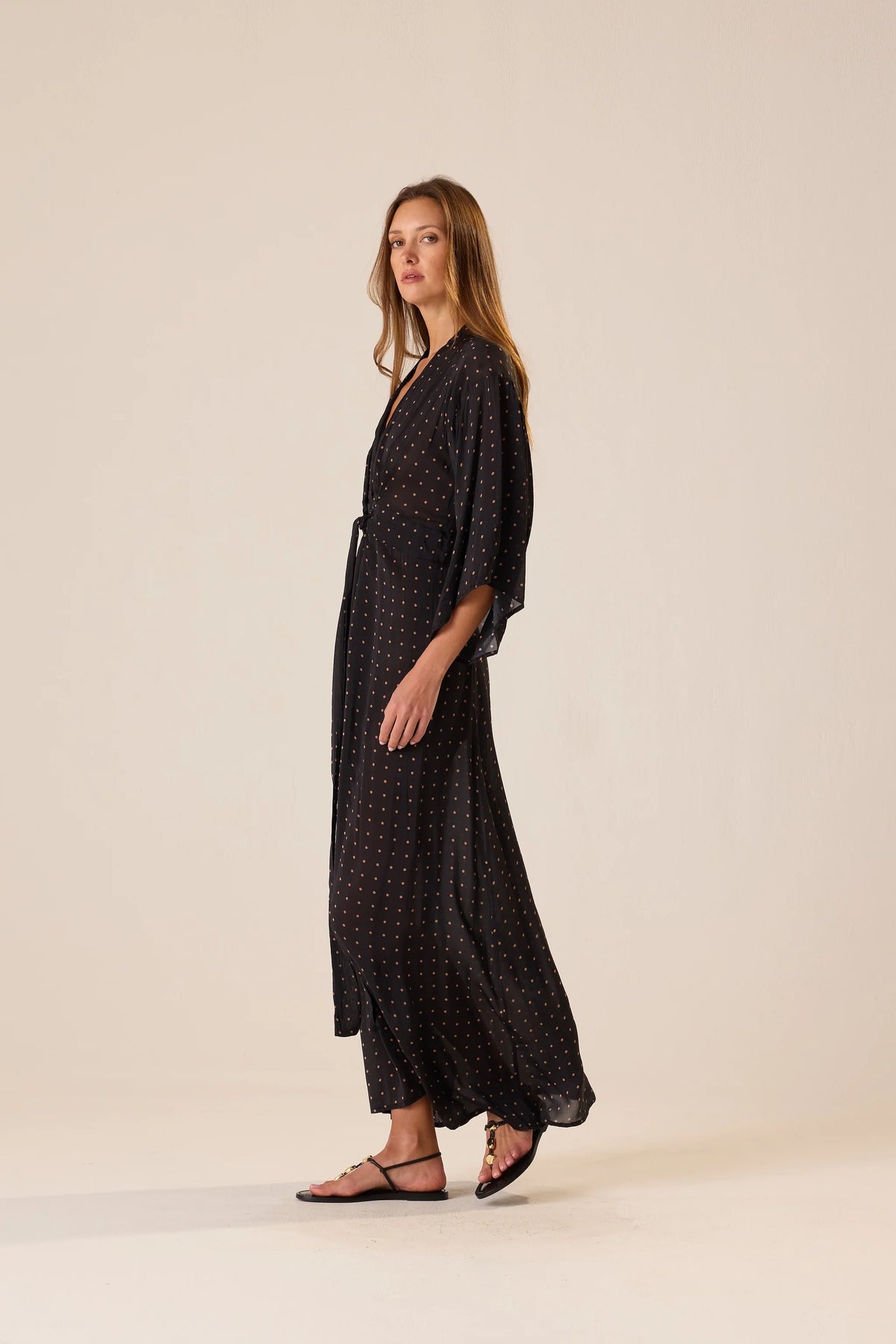Natalie Martin Sarah Dress in black with polka dots, shown sideways with sandals, minimal backdrop.