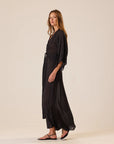 Natalie Martin Sarah Dress in black with polka dots, shown sideways with sandals, minimal backdrop.