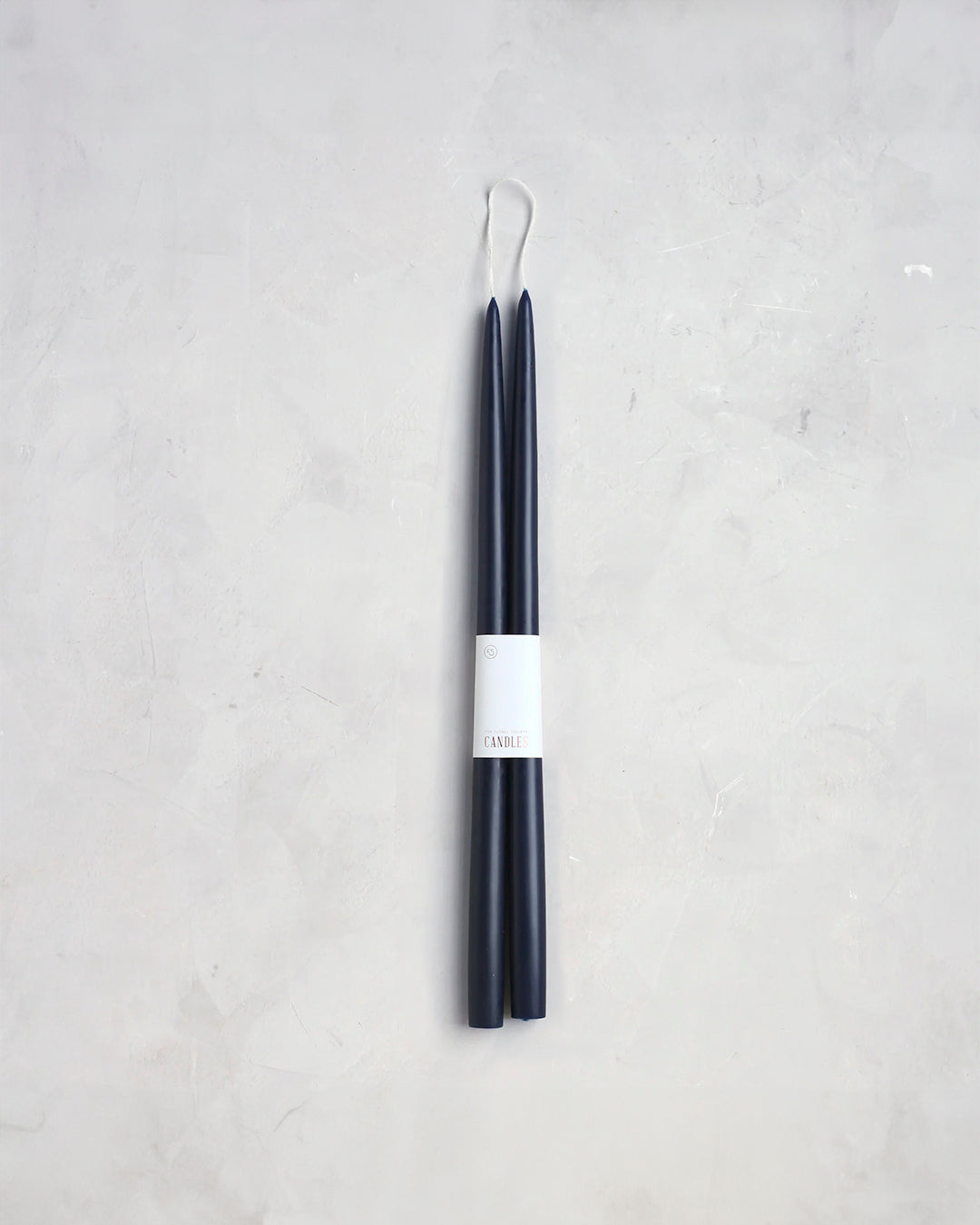 A sophisticated pair of The Floral Society's 18" Taper Candles, presented in a dark blue hue and designed with sleek, slim lines. Each candle features a white label reading "CANDLE," complemented by an elegant loop of white thread at the top. Set against a light gray, textured background, they emanate a graceful ambiance perfect for any setting.