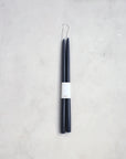 A sophisticated pair of The Floral Society's 18" Taper Candles, presented in a dark blue hue and designed with sleek, slim lines. Each candle features a white label reading "CANDLE," complemented by an elegant loop of white thread at the top. Set against a light gray, textured background, they emanate a graceful ambiance perfect for any setting.