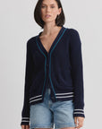 A woman with a bob haircut wears the Chelsea Cardigan by One Grey Day, featuring striped tipping and a half-cardigan stitch, over denim shorts against a plain light background.