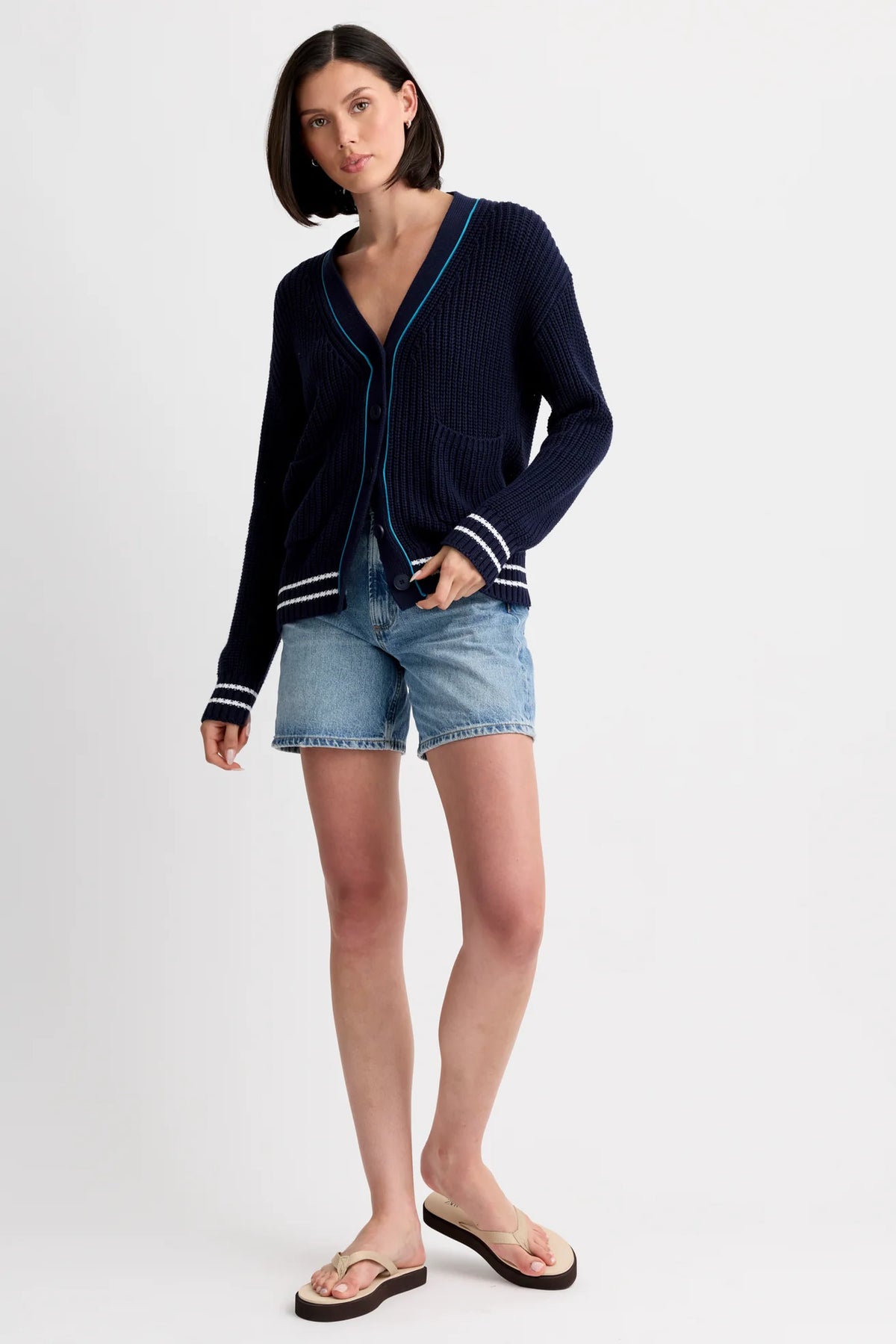 A woman with short dark hair and a neutral expression stands against a plain background wearing the One Grey Day Chelsea Cardigan in navy blue with white sleeve stripes, paired with denim shorts and beige flip-flops.