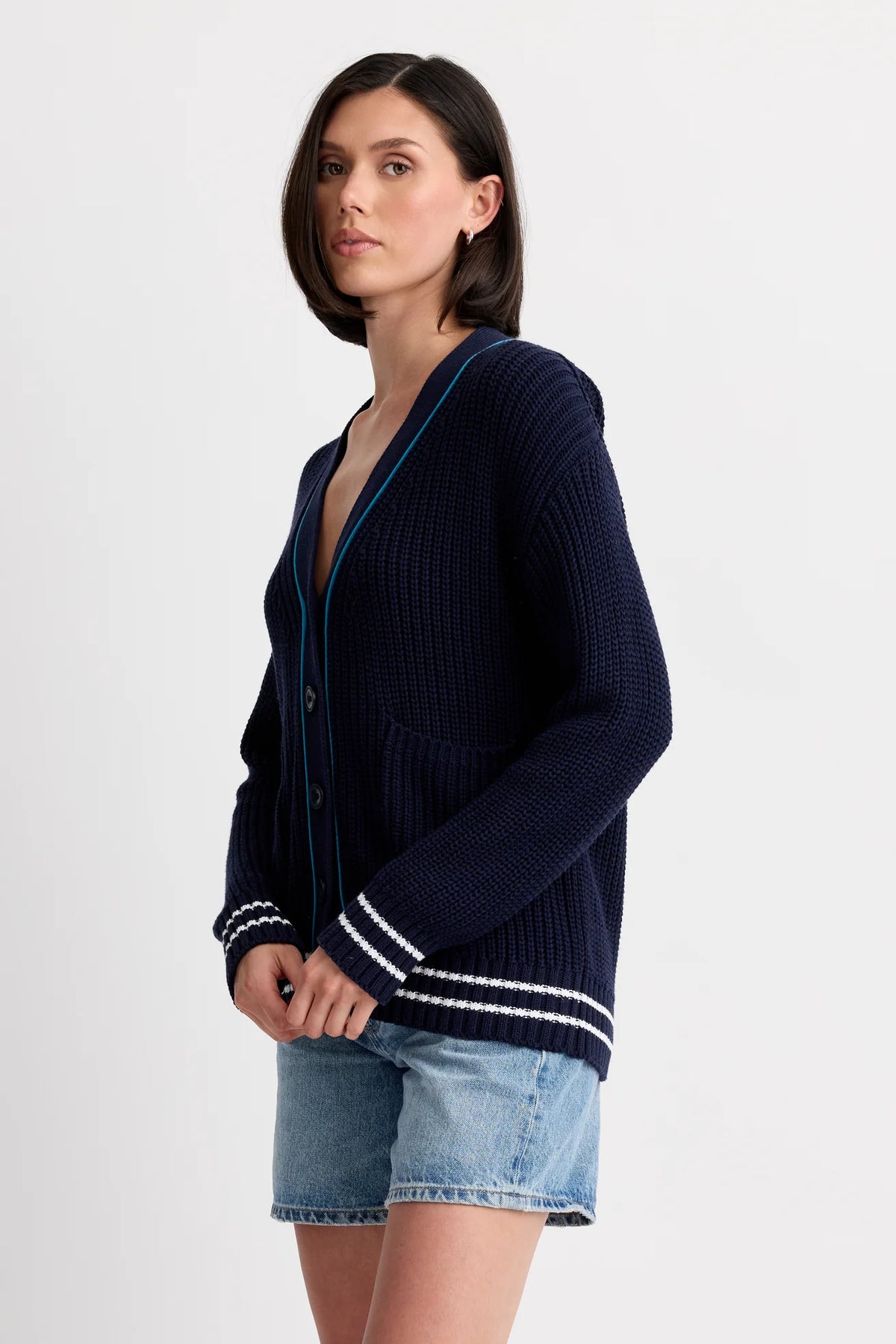A person wears the One Grey Day Chelsea Cardigan, a women's navy knit cardigan with white and blue trim, paired with light blue denim shorts, standing against a plain white background.
