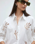 A woman wears the Loula Blouse by SUNCOO—a white button-up with lace cutouts made from 100% organic cotton—paired with oversized square sunglasses and pearl earrings, standing against a plain background.