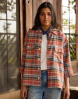 A woman with long dark hair stands outdoors by wooden doors, wearing a red plaid shirt over a white tee, blue jeans, and The HG Bandana by Hey Gang around her neck. She has a neutral expression.