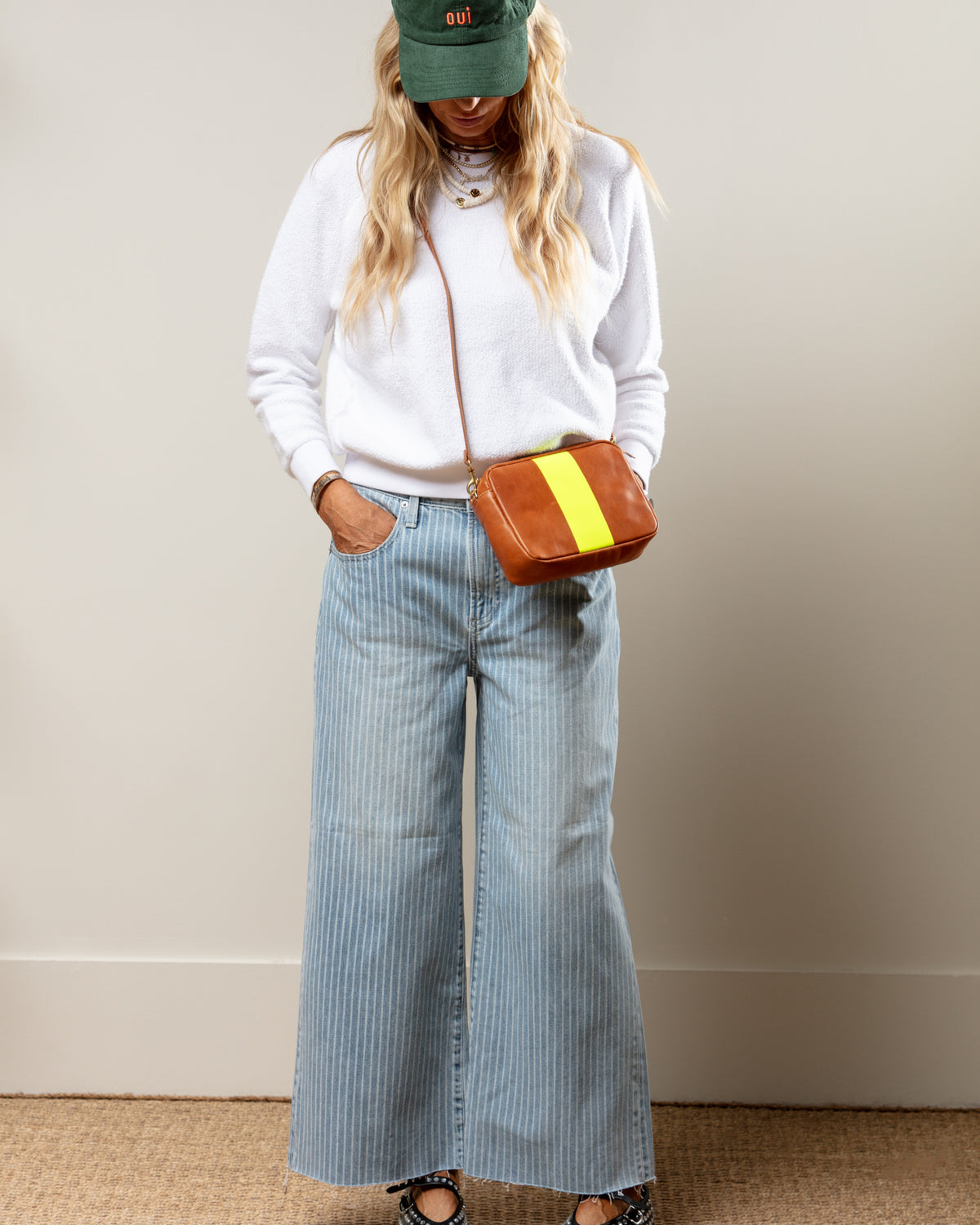 Woman models striped wide-leg jeans and white sweater, styled with green hat and crossbody bag for a relaxed look.