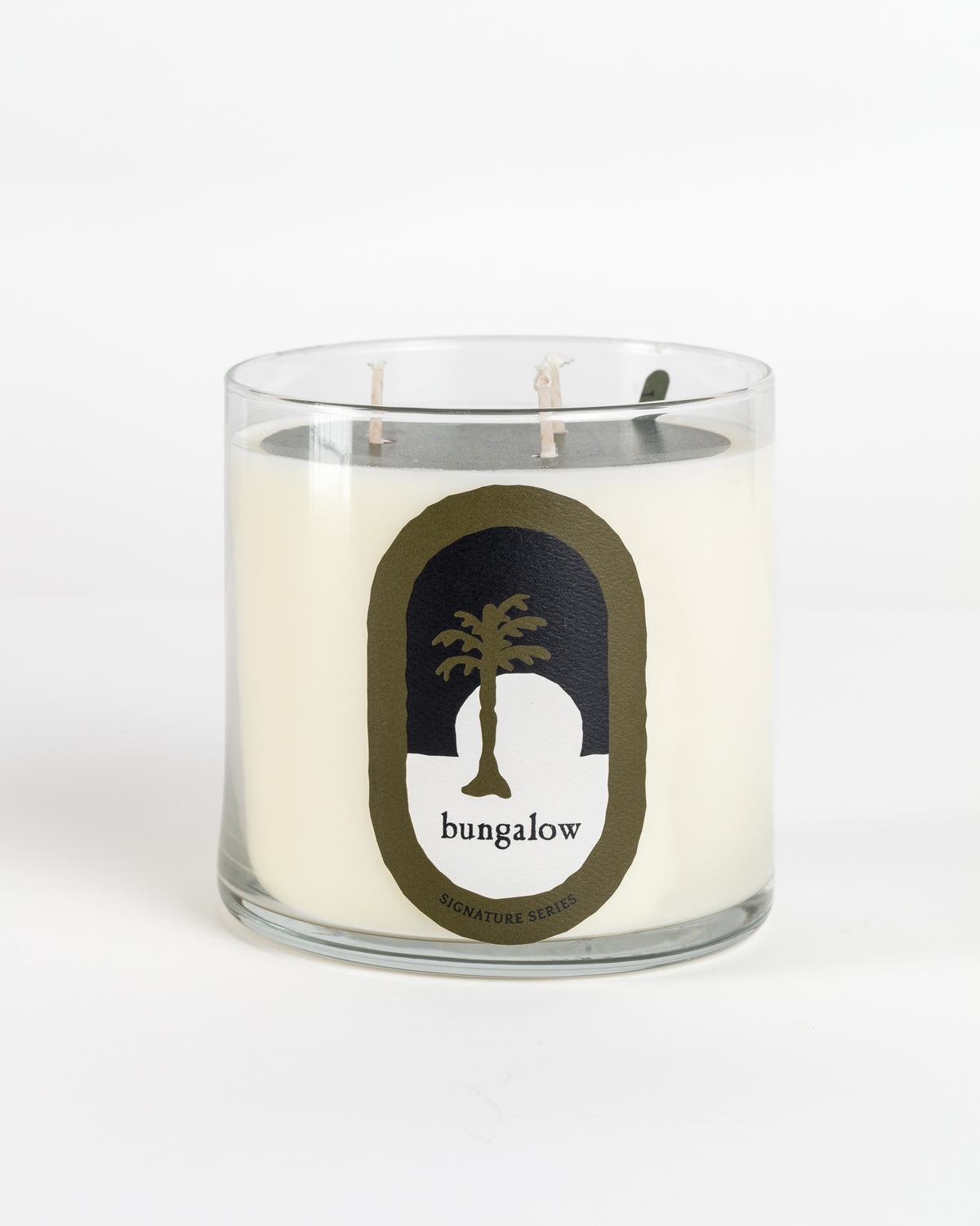 The New Signature Candle by Mame, infused with sandalwood, features a white candle in a glass jar with two wicks. Its label showcases a black and white design with a palm tree silhouette and the word "bungalow" in a simple font, all set against a plain white background.