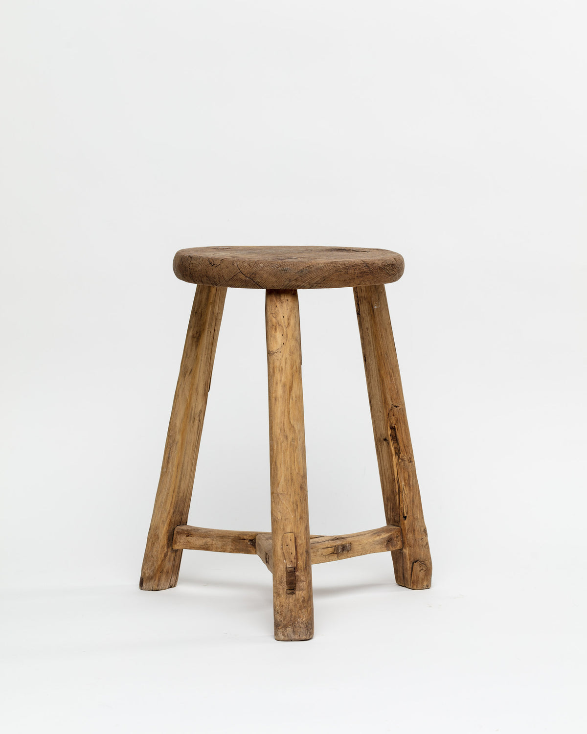 The ROUND STOOL by Indus Design Imports showcases a rustic wooden design featuring a round seat and four sturdy legs. This stool exudes timeless elegance with its natural, unfinished appearance that beautifully highlights the wood's texture and grain. The artisan craftsmanship is evident in the horizontal braces connecting the legs, providing added stability. Photographed against a plain white background, this piece stands out for its simplicity and charm.