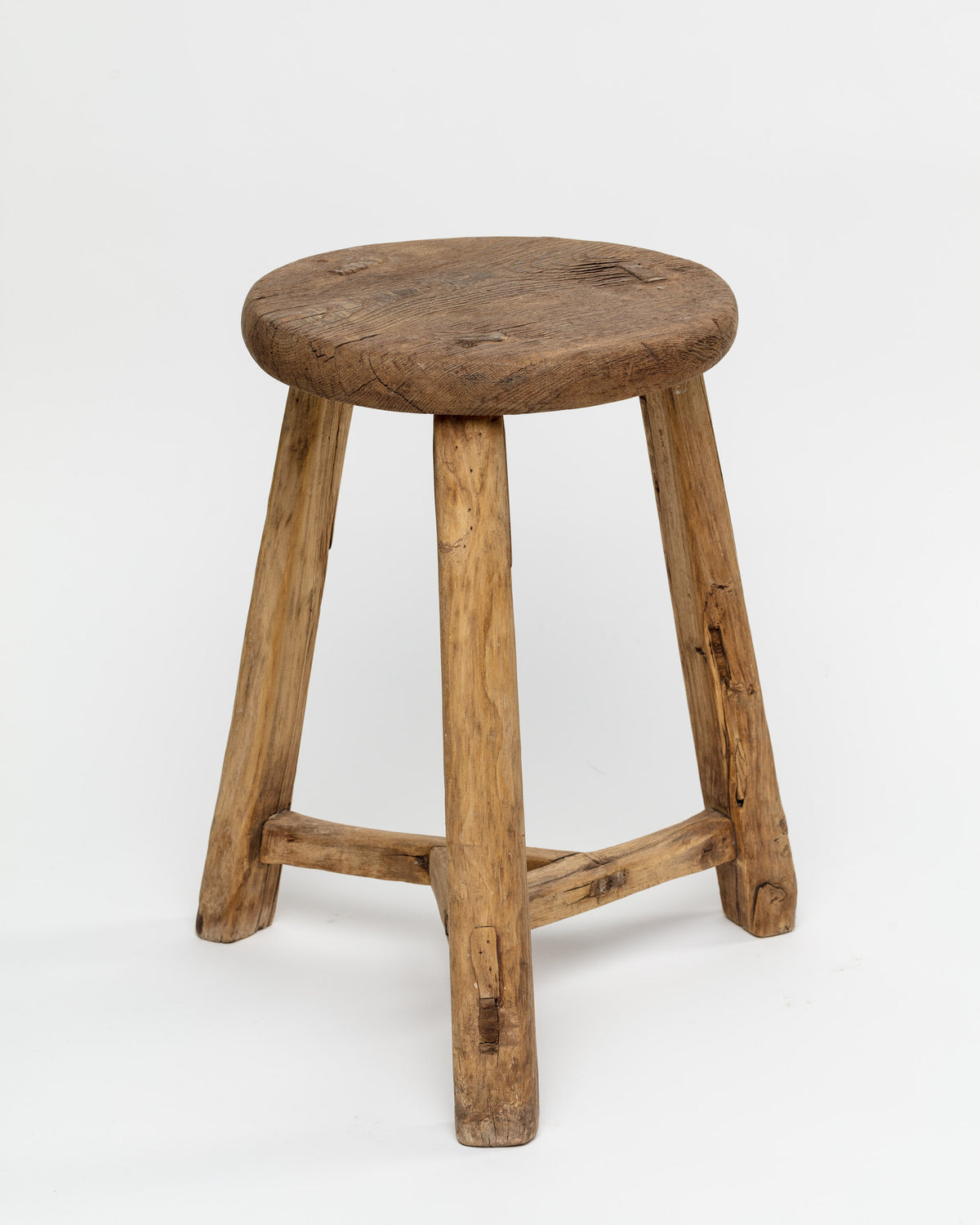 The ROUND STOOL by Indus Design Imports features a rustic wooden design with a round, worn seat and three slightly angled, sturdy legs connected by a triangular support structure. The stool's visible wood grain and imperfections showcase artisan craftsmanship, giving it a timeless, handmade appearance against a plain white background.