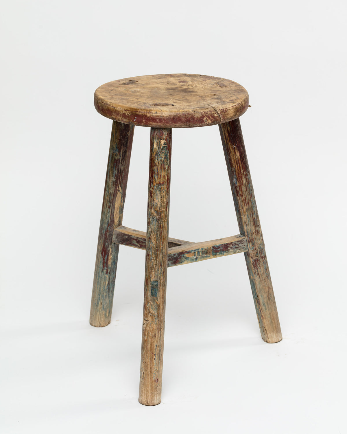 The ROUND STOOL by Indus Design Imports is a rustic, weathered wooden stool featuring a round seat and three legs. Showcasing timeless elegance, the stool is aged with worn paint and visible signs of use, reflecting its vintage charm. This piece of artisan craftsmanship stands out beautifully against a plain white background.