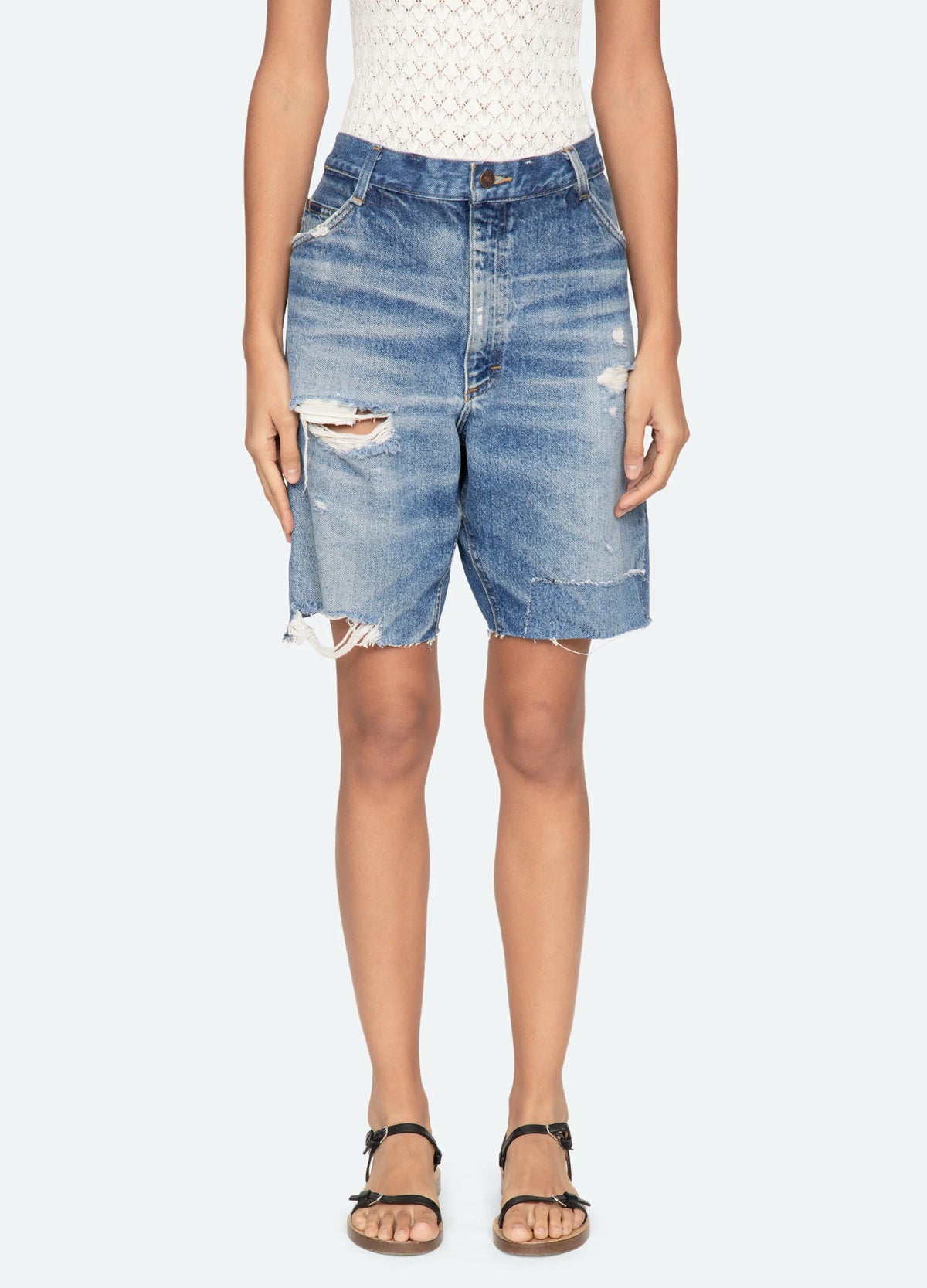 A person wears a sleeveless patterned top and Sea New York’s Milene Denim Knee Short, featuring a relaxed fit and frayed hems, paired with black sandals against a plain light background.