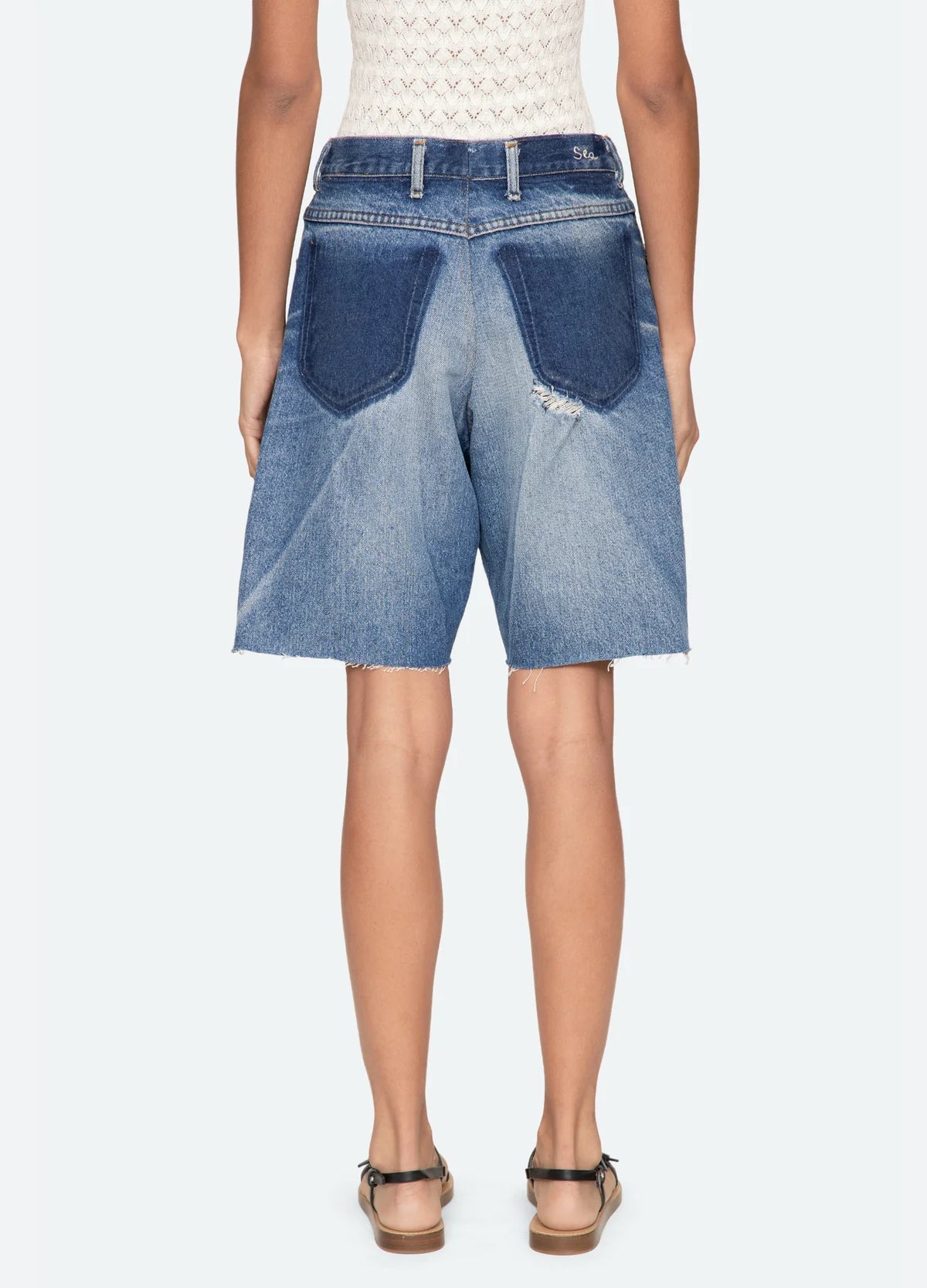 A person is seen from behind wearing Sea New York’s Milene Denim Knee Short—relaxed fit, high-waisted, vintage-inspired with large dark blue back pockets and frayed hems—styled with sandals and a sleeveless patterned top.