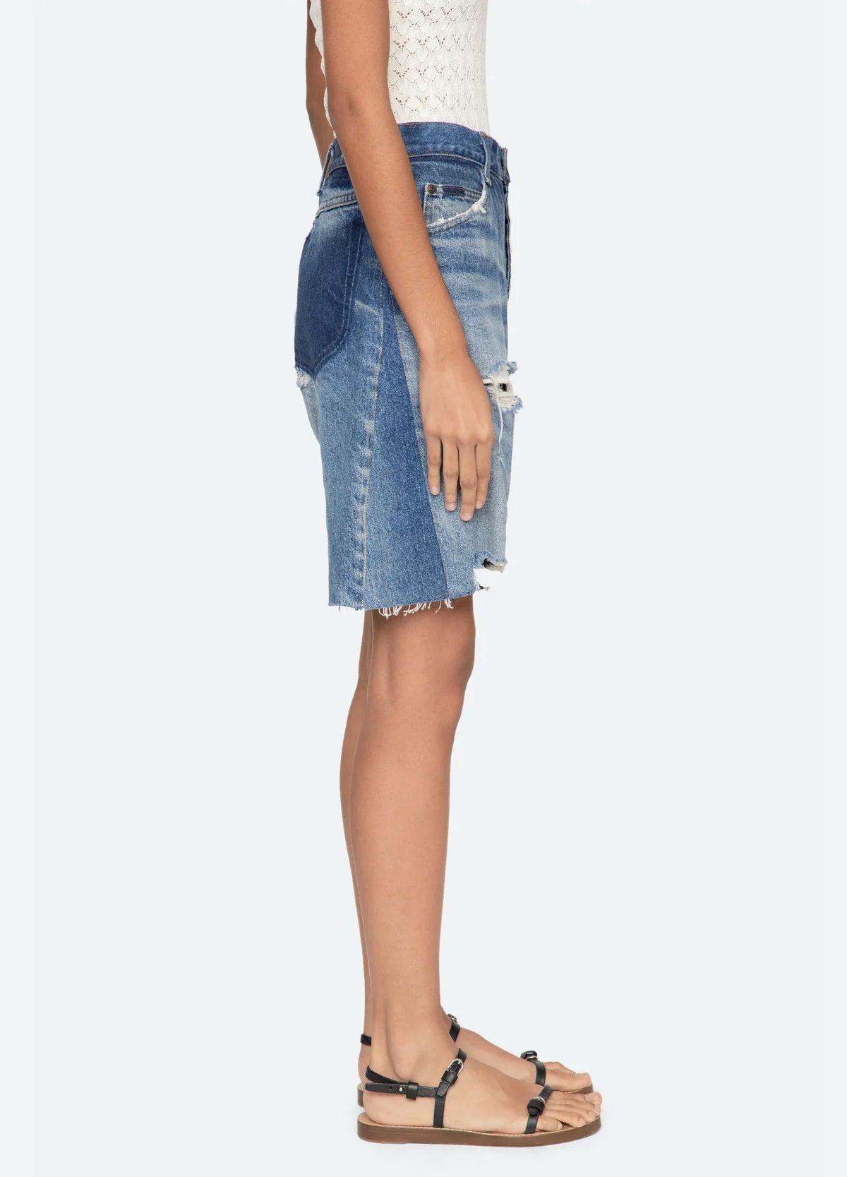 A woman stands in profile wearing a sleeveless white top, tan flat sandals with black straps, and the Milene Denim Knee Short by Sea New York—a knee-length vintage patchwork skirt featuring frayed hem and distressed details.