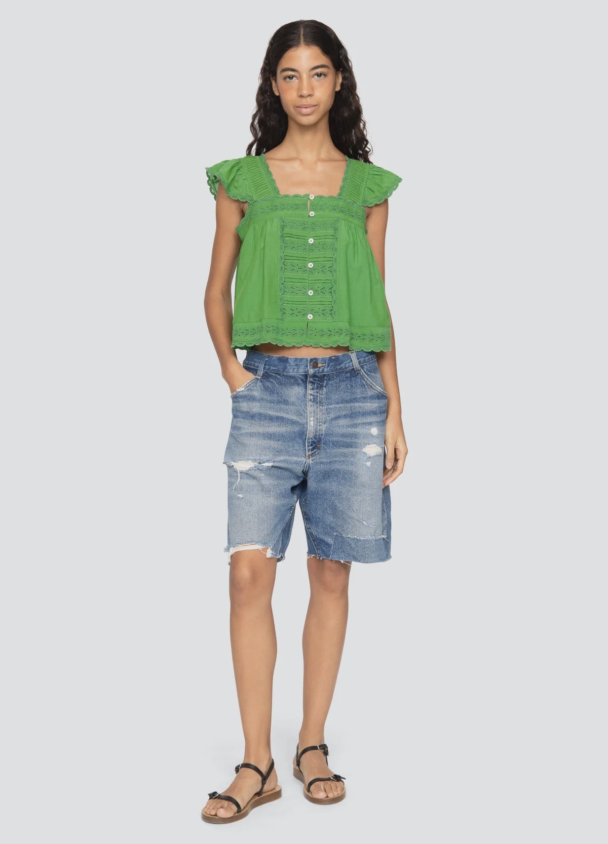 A woman stands facing forward in a bright green button-up blouse, Milene Denim Knee Shorts by Sea New York, and black sandals. She has long wavy hair and a relaxed expression.