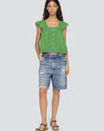 A woman stands facing forward in a bright green button-up blouse, Milene Denim Knee Shorts by Sea New York, and black sandals. She has long wavy hair and a relaxed expression.