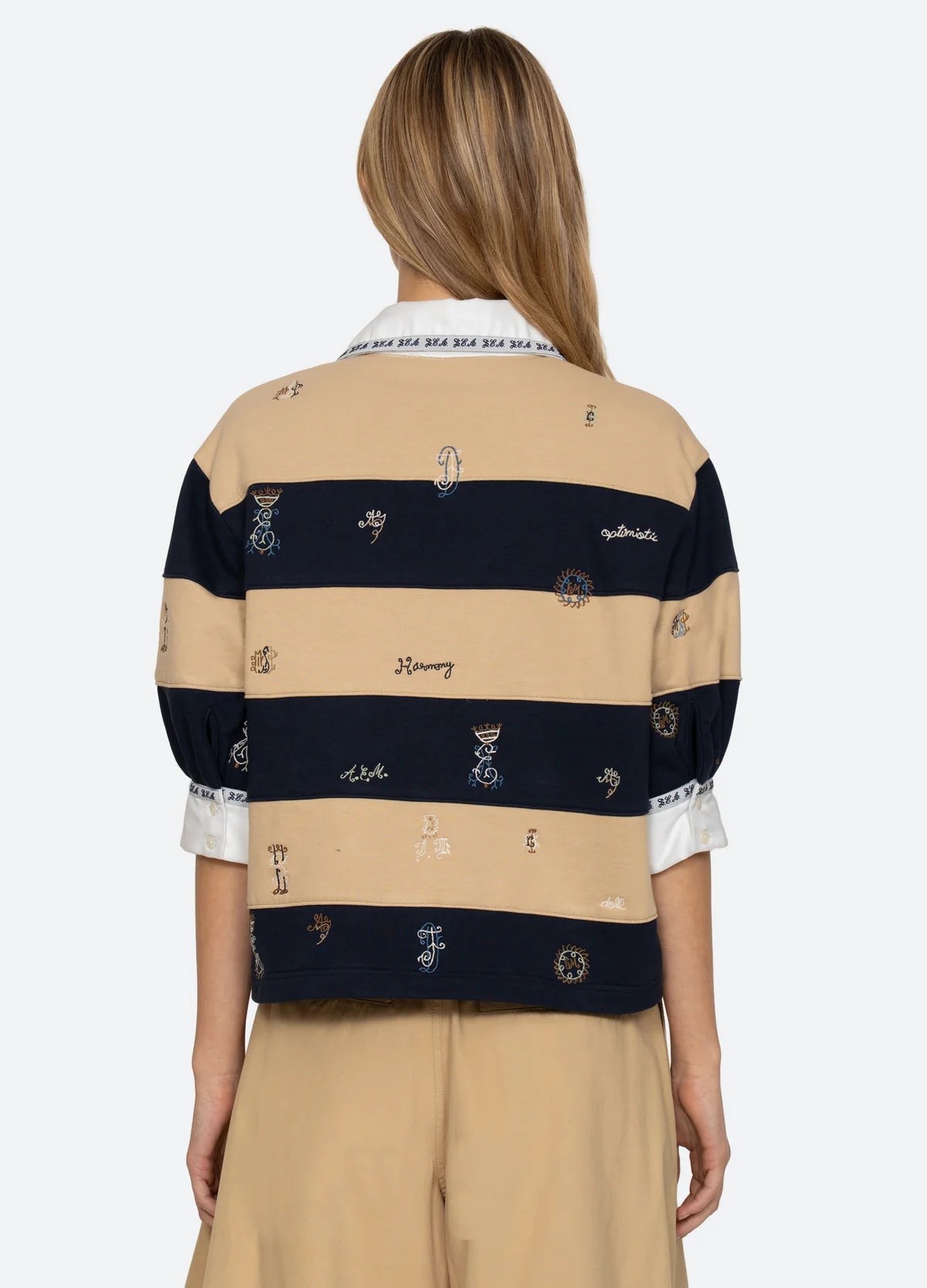 A woman with long, light brown hair wears the Sea New York Juliet SS Rugby—a loose, embroidered top with puff sleeves—layered over a white collared shirt. She faces away, showcasing this chic workwear-inspired style.