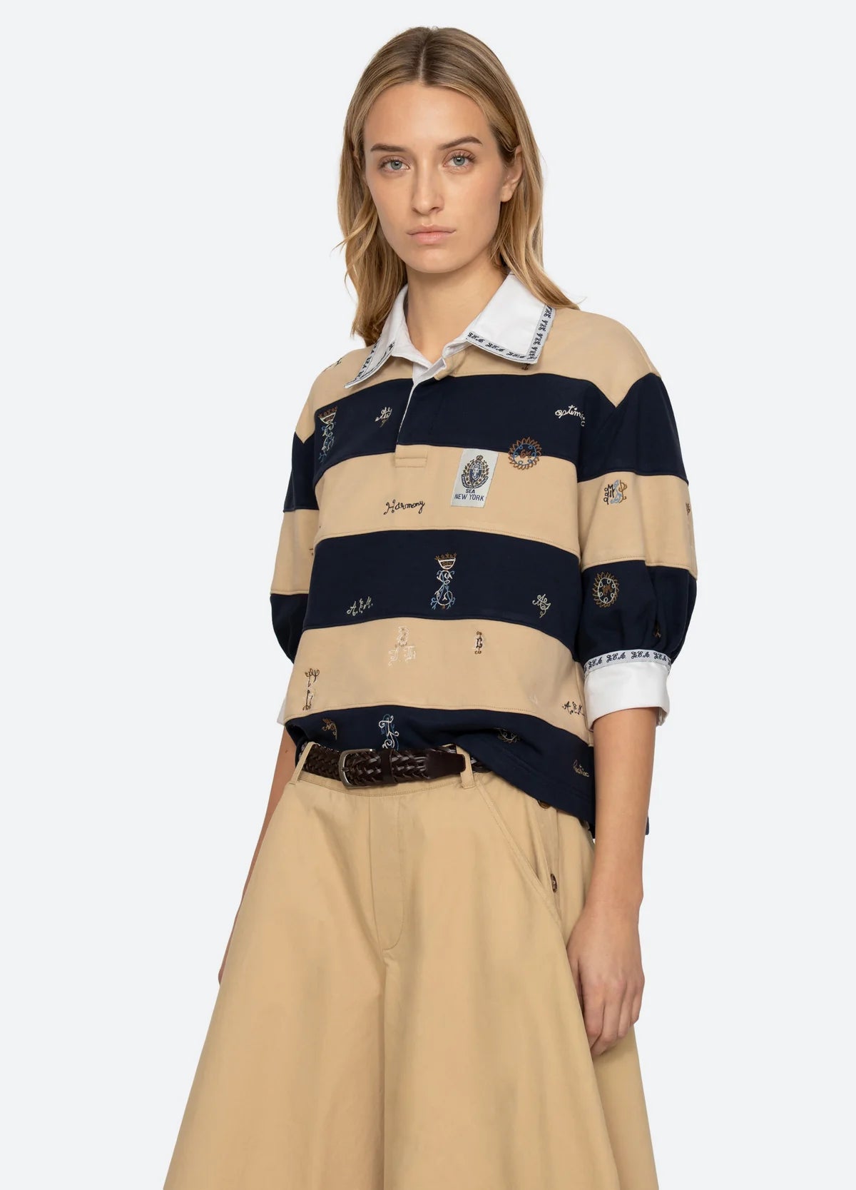 A woman wears the Sea New York Juliet SS Rugby—a puff-sleeve top with beige and navy stripes plus a white collar—paired with a beige skirt and braided brown belt, standing against a plain light background.