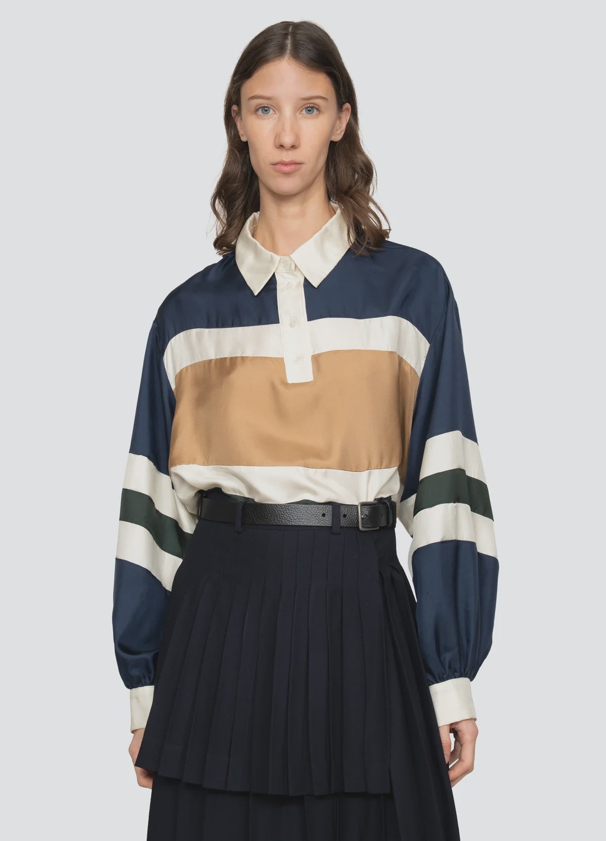 A woman wears the Sea New York Juliet Polo, featuring bold navy, gold, cream, and green stripes, paired with a high-waisted pleated black skirt and belt. She stands against a plain light background.