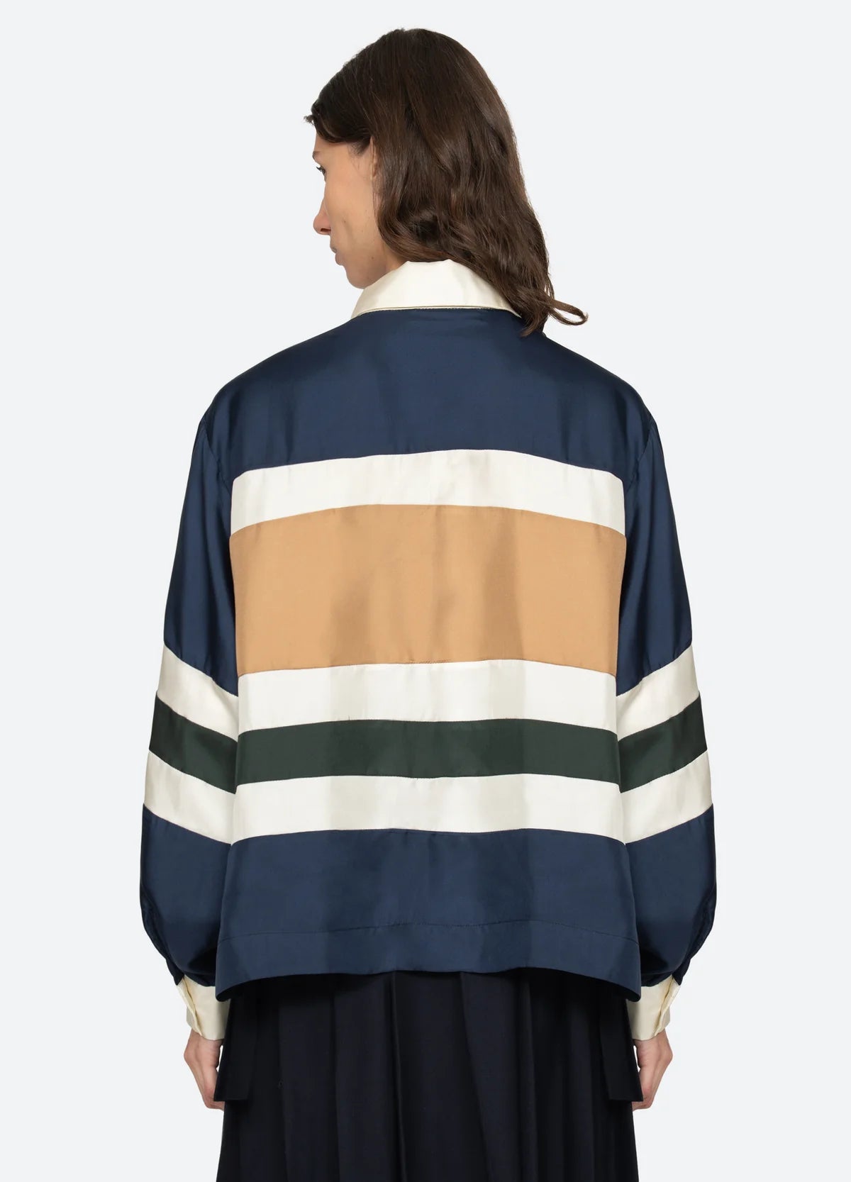 A person with shoulder-length brown hair is seen from the back wearing the Sea New York Juliet Polo, a loose modern-fit jacket with wide navy, tan, white, and dark green stripes and voluminous sleeves, against a plain light background.