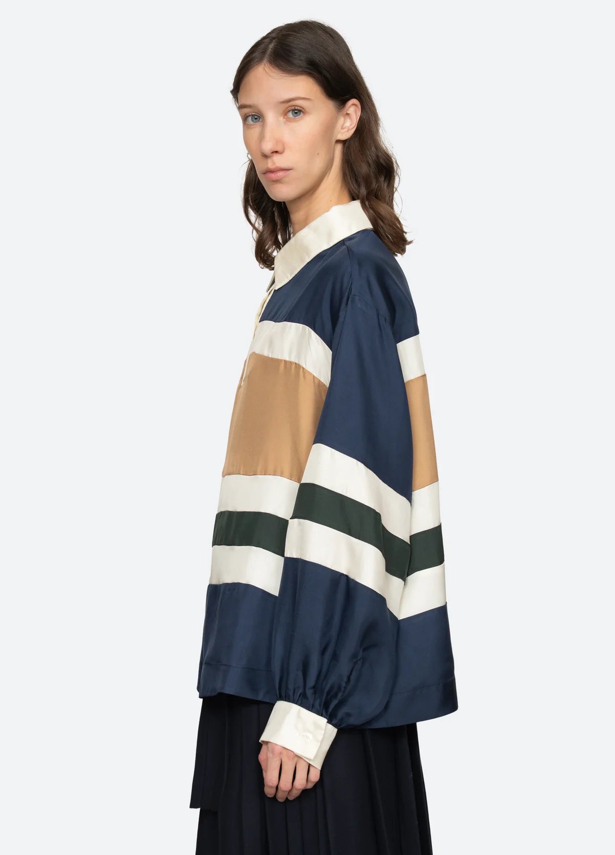 A woman with wavy brown hair models the Sea New York Juliet Polo, featuring voluminous sleeves and horizontal stripes in navy, tan, cream, and green. She pairs it with a black skirt and stands sideways against a light background.