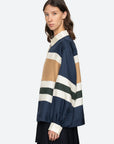 A woman with wavy brown hair models the Sea New York Juliet Polo, featuring voluminous sleeves and horizontal stripes in navy, tan, cream, and green. She pairs it with a black skirt and stands sideways against a light background.