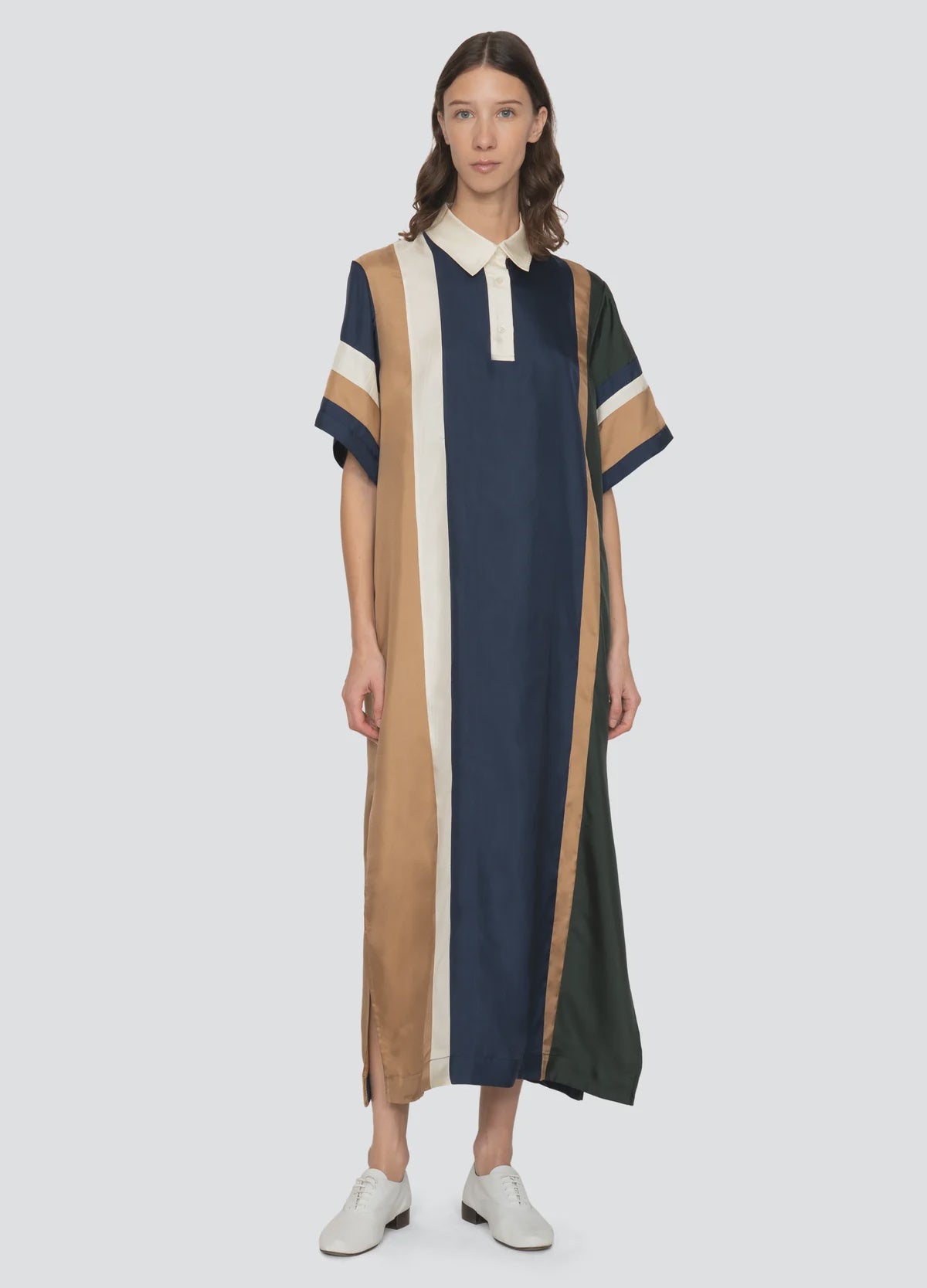 A person wears the Sea New York Juliet Polo Maxi Dress—an ankle-length, loose-fit dress with navy, tan, cream, and dark green vertical stripes, white collar, short sleeves with tan and green bands—and white shoes against a plain background.