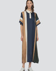 A person wears the Sea New York Juliet Polo Maxi Dress—an ankle-length, loose-fit dress with navy, tan, cream, and dark green vertical stripes, white collar, short sleeves with tan and green bands—and white shoes against a plain background.