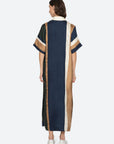 A person with short brown hair wears white shoes and the Sea New York Juliet Polo Maxi Dress—color-blocked with vertical navy, cream, and brown stripes and matching short sleeves—while facing away.