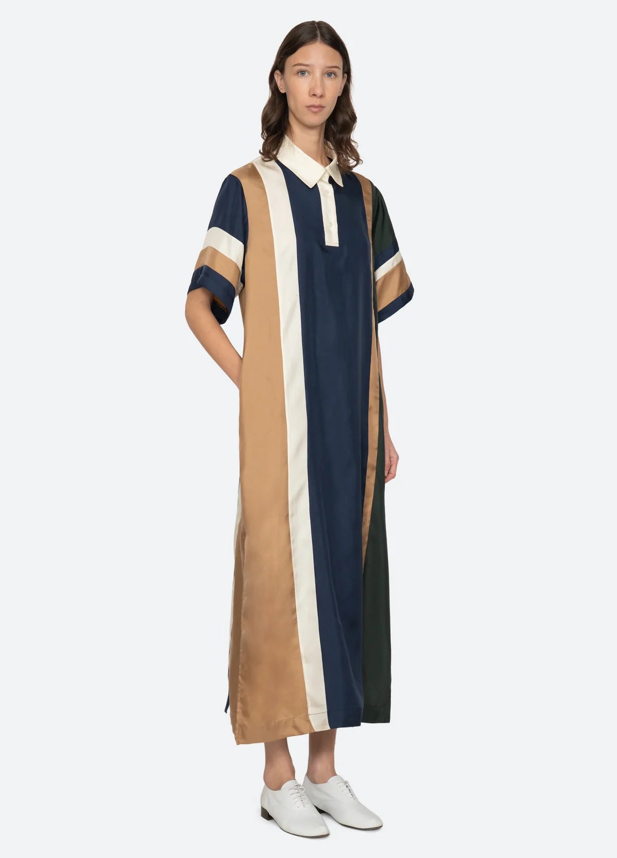 A woman wears the Sea New York Juliet Polo Maxi Dress, a loose-fitting, short-sleeve style with vertical navy, tan, cream, and green stripes. She stands with one hand in her pocket and white shoes against a plain background.