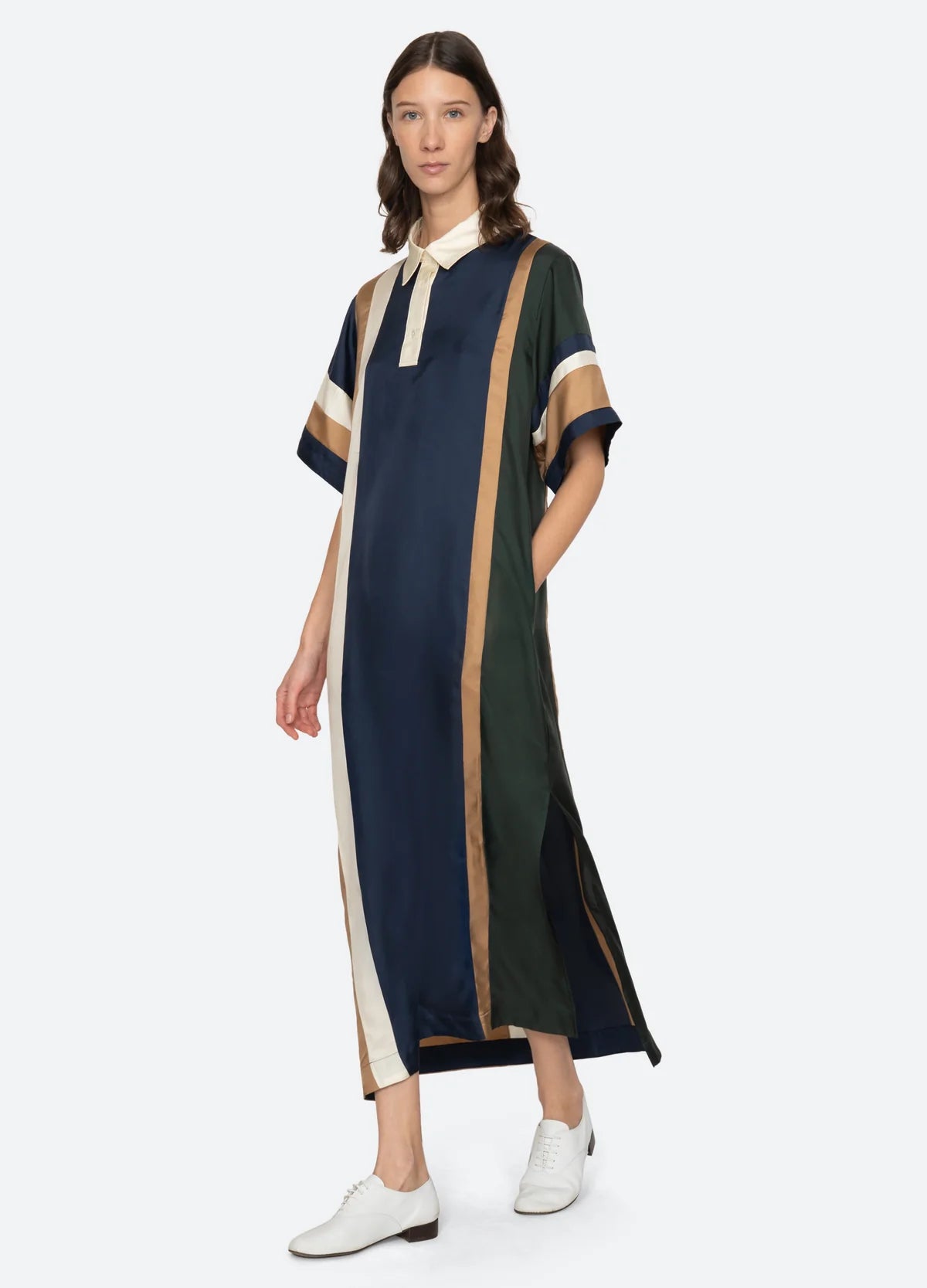 A woman wears the Sea New York Juliet Polo Maxi Dress— a loose, short-sleeved style with vertical color-block stripes in navy blue, green, beige, and gold—paired with classic white oxford shoes.
